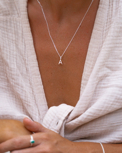 A Buchstabenkette aus silber um den Nacken eines weiblichen Models. Sie trägt Leinenhemd, silberringe, sonstigen Schmuck und ist gebräunt.