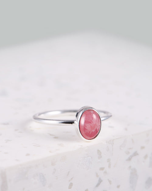 Auf dem Bild liegt ein Ring auf einer weißen Marmorplatte. Der Ring hat ein schmales rundes Band, oben ist ein rosafarbener Rhodochrosit-Stein eingefasst. Der Stein schimmert in verschiedenen Rosa-Tönen.