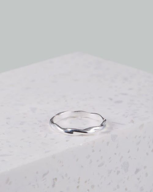 Auf dem Bild ist ein weißer Marmorklotz zu sehen, auf dem ein silberner Ring liegt. Der Ring ist in sich eingedreht. Der Ring bekommt dadurch eine wellenähnliche Optik. Das Silber glänzt und reflektiert.