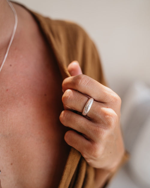 Auf dem Bild ist eine Hand zu sehen, die den Rand eines braunen Oberteils festhält. Die Frau trägt einen Ring von Verlan Jewellery in wasserfestem Sterling Silber am Mittelfinger. Der Ring ist etwas breiter und die Oberfläche ist nach außen gewölbt. Auf dem Ring sind feine Linien quer eingraviert, die in schmalem Abstand zueinander stehen.