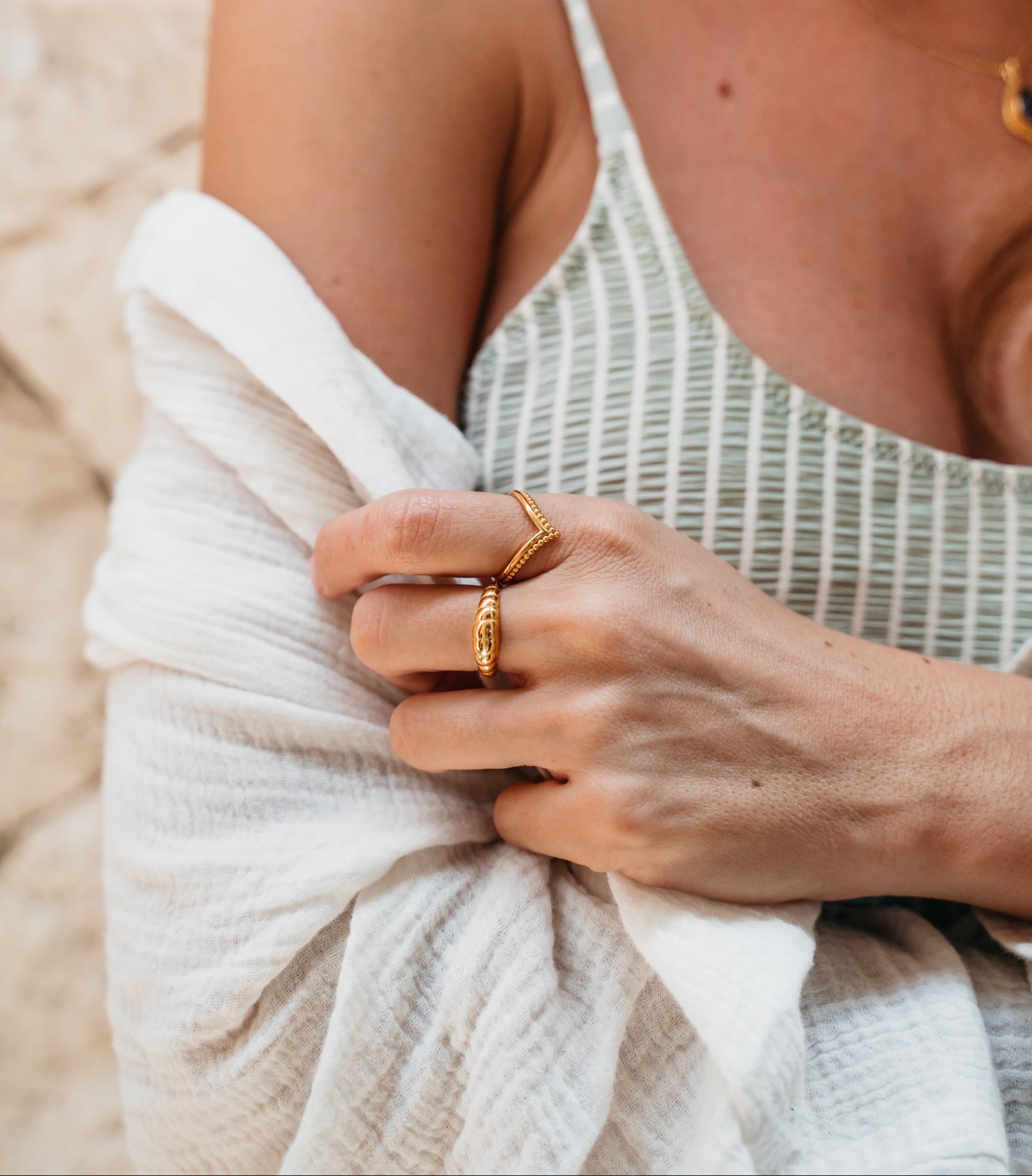 Goldene Ringe an weiblicher Model Hand. Ein V Ring mit Perlen im Boho Vibe und ein Croissant Ring in Gold. Handgefertigte Schmuckstücke aus Bali. Das Model trägt ein gestreiftes Top und eine weiße Musselin Bluse.