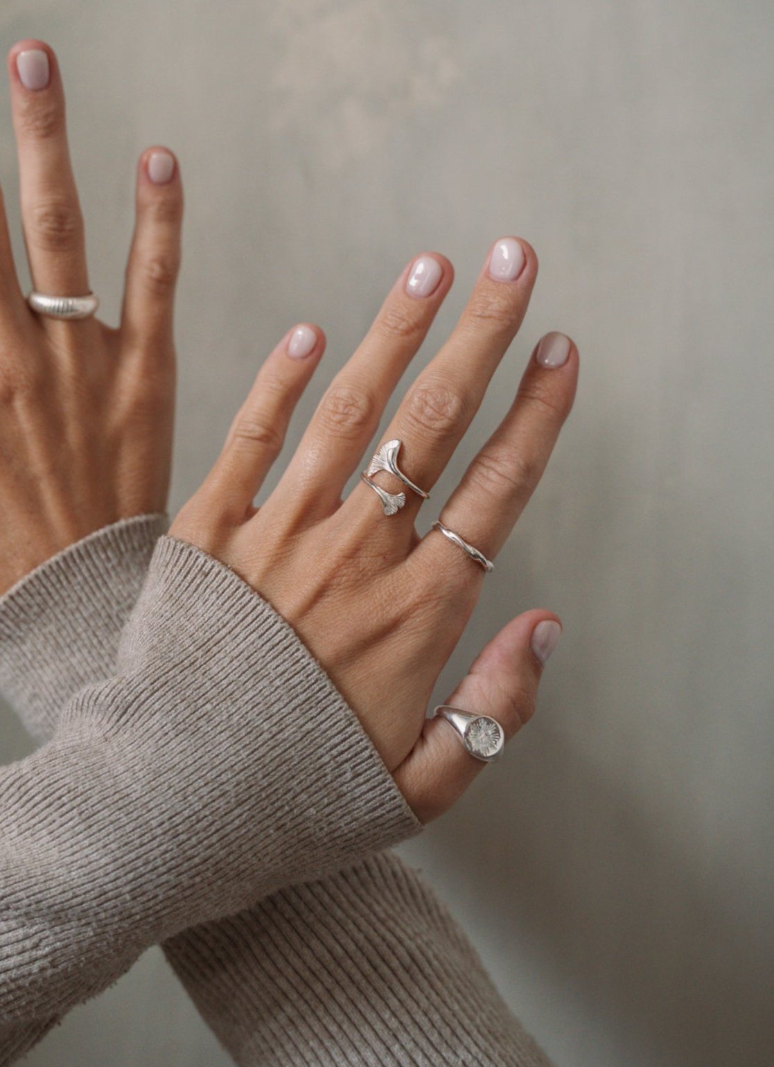 Auf dem Bild überkreuzen sich zwei Hände. Das Model trägt weißen Nagellack und man sieht Ärmel eines grauen Strickpullis. Der Hintergrund ist eine graue Betonwand. Das Model trägt drei silberne Ringe von Verlan Jewellery an ihrer linken Hand und einen an der Rechten. Sie trägt an der linken Hand am Mittelfinger einen Ring mit zwei Ginkgoblättern, den eingedrehten Legian-Ring am Zeigefinger und den Siegelring-ähnlichen Medewi Ring am Daumen. Am rechten Ringfinger trägt sie den etwas breiteren Kedungu Ring. 