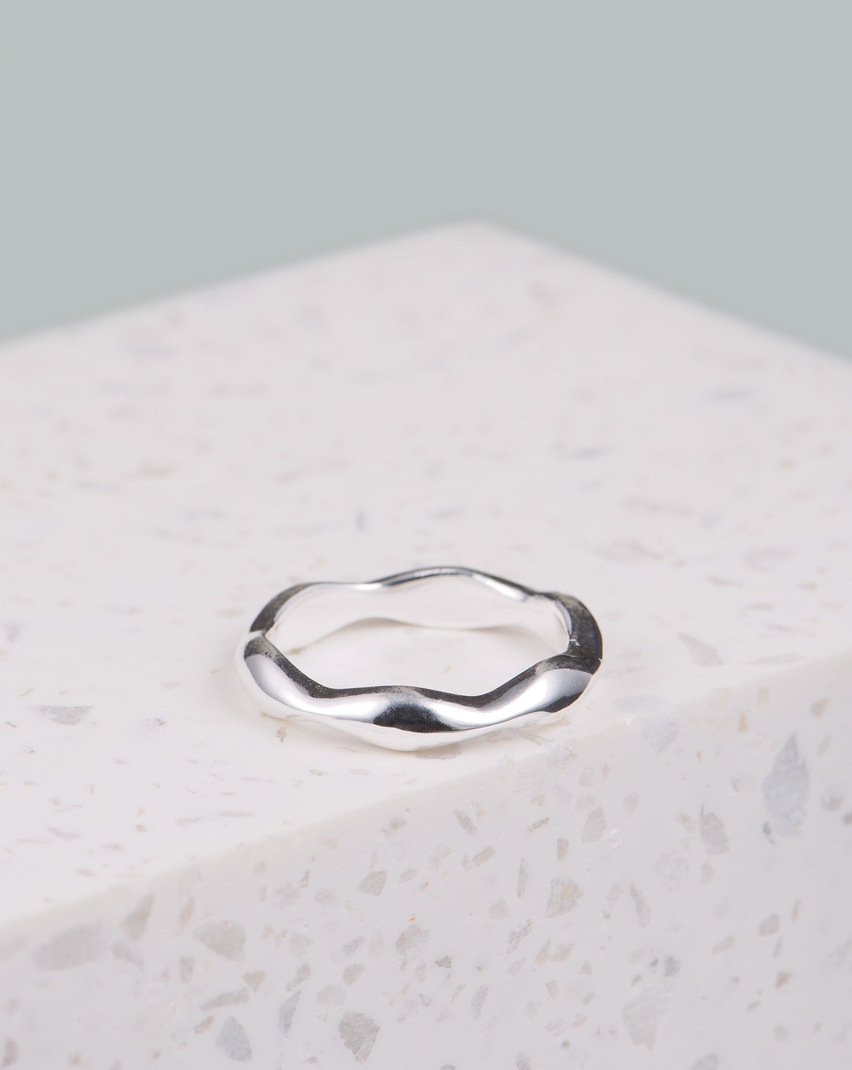 Der auf Bali handgefertigte Melambai Ring liegt hier auf einem weißen Marmorblock in der Mitte des Bildes. Der Hintergrund ist etwas verschwommen, das Foto ist hell. Der Ring glänzt und wird abwechselnd dicker und dünner, es sieht aus wie eine Welle. 