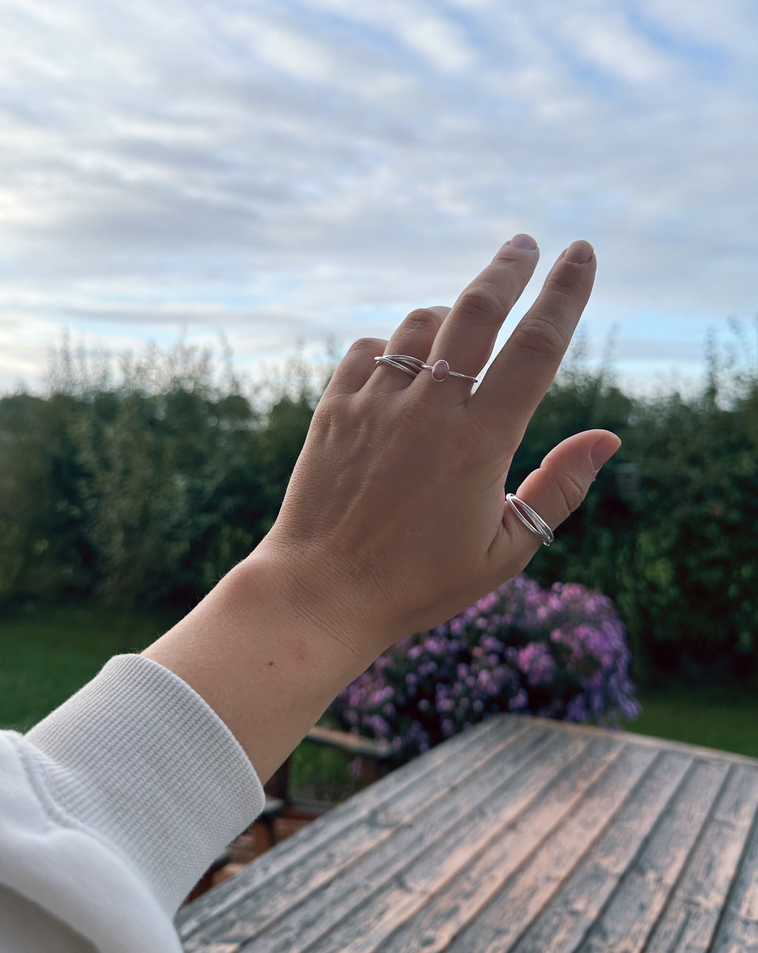 Das Bild zeigt eine Hand, die in Richtung Himmel gestreckt ist. Im Hintergrund erkennt man einen Garten mit grüner Hecke und einem Busch mit lilafarbenen Blüten. An der Hand trägt das Model drei Silberringe von Verlan Jewellery. Am Ringfinger den Tiga Ring, welcher aus drei einzelnen Ringen besteht und somit in sich beweglich ist, am Mittelfinger einen Silberring mit rosafarbenen Rhodochrosit und am Daumen ebenfalls den Tiga Antistressring in einer größeren Größe. 