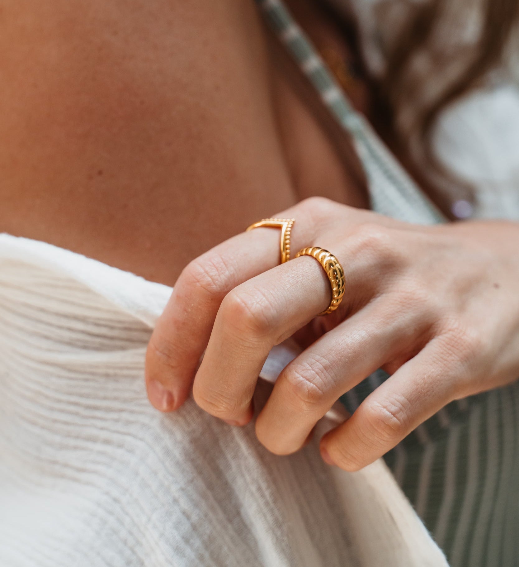 Goldene Ringe an weiblicher Model Hand. Ein V Ring mit Perlen im Boho Vibe und ein Croissant Ring in Gold. Handgefertigte Schmuckstücke aus Bali. Das Model trägt ein gestreiftes Top und eine weiße Musselin Bluse.