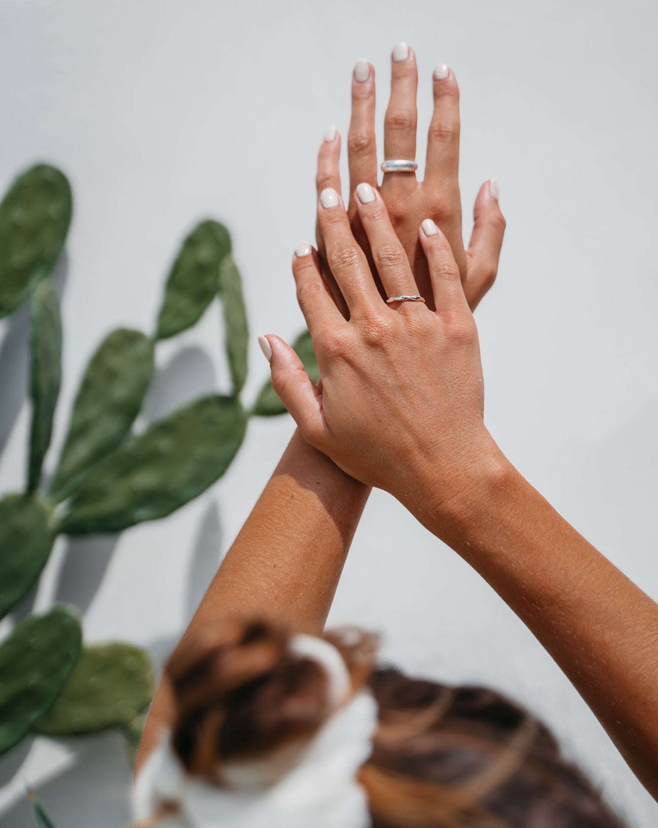 Das Bild zeigt eine Frau, die beide Hände übereinander gelegt in die Luft streckt. Die rechte Hand liegt über der Linken und verdeckt diese etwas. Die Frau trägt weißen Nagellack. An ihrem rechten Ringfinger trägt sie den Legian-Ring in silber, welcher eine eingedrehte Optik hat. An ihrem linken Mittelfinger ist ein breiterer, dickerer Silberring zu sehen. Im Hintergrund erkennt man etwas unscharf einen grünen Kaktus.