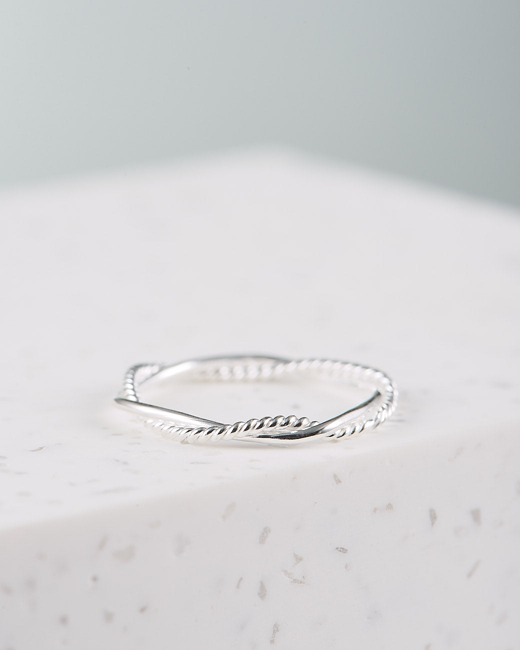 Auf dem Foto sieht man den Sari Ring in Silber. Er liegt auf einem weißen Marmorblock. Der Hintergrund ist grau und unscharf. Der auf Bali handgefertigte Ring besteht aus zwei dünnen Bändern, die locker ineinander verdreht sind. ein Band ist glatt, das andere ist eng spiralförmig eingedreht. 