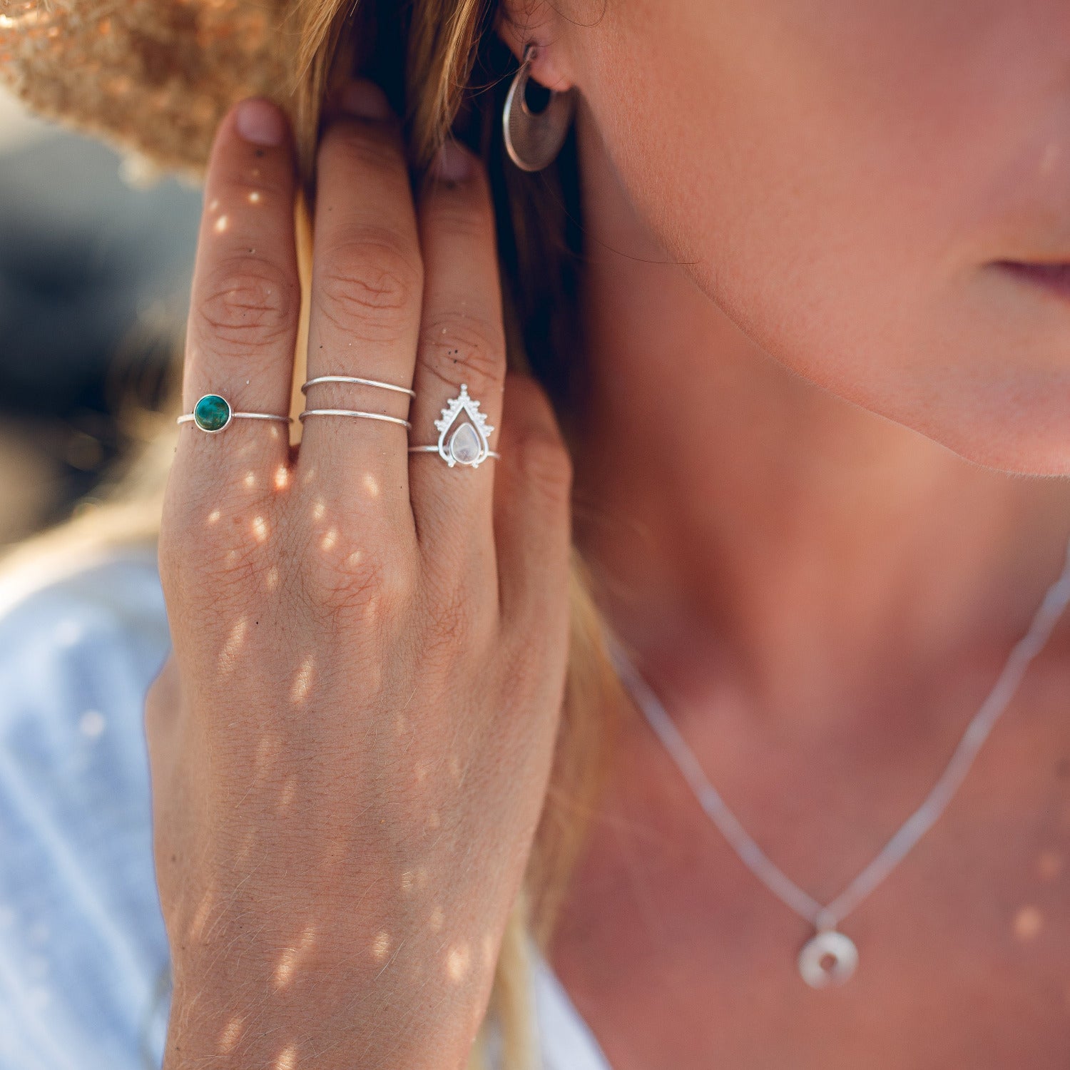 Auf dem Bild sieht man die Hand und den Teil des Kopfes eines Models. Auf der Hand sind Reflektionen der Sonne zu sehen, welche von einem Strohhut stammen. Das Model trägt wasserfesten Silberschmuck von Verlan Jewellery: Die Nusa Dua Silberkette mit Mond Anhänger, den Lovina Silberohrring und drei Ringe. Am Ringfinger den Manta Ring mit einem Mondstein und in Ornamente eingefasst, am Mittelfinger den Binging Ring, am Zeigefinger den Ubud Ring mit einem runden Türkis Stein. 