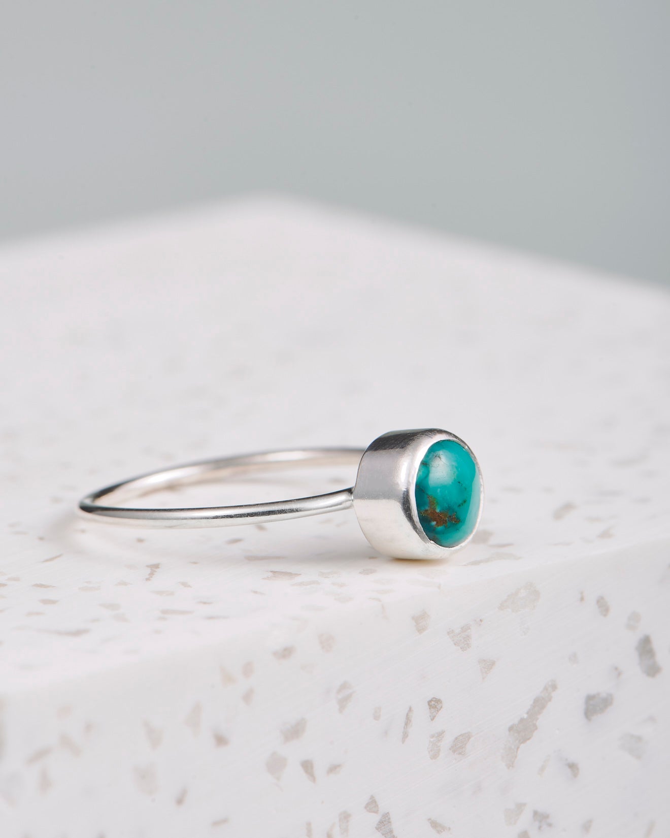 Ein Silberring mit türkisfarbenem Stein liegt auf einer weißen Marmorplatte. Der Stein ist rund und in Silber eingefasst. Der Hintergrund des Bildes ist grau und verschwommen. Der Ubud Ring liegt im Fokus. 