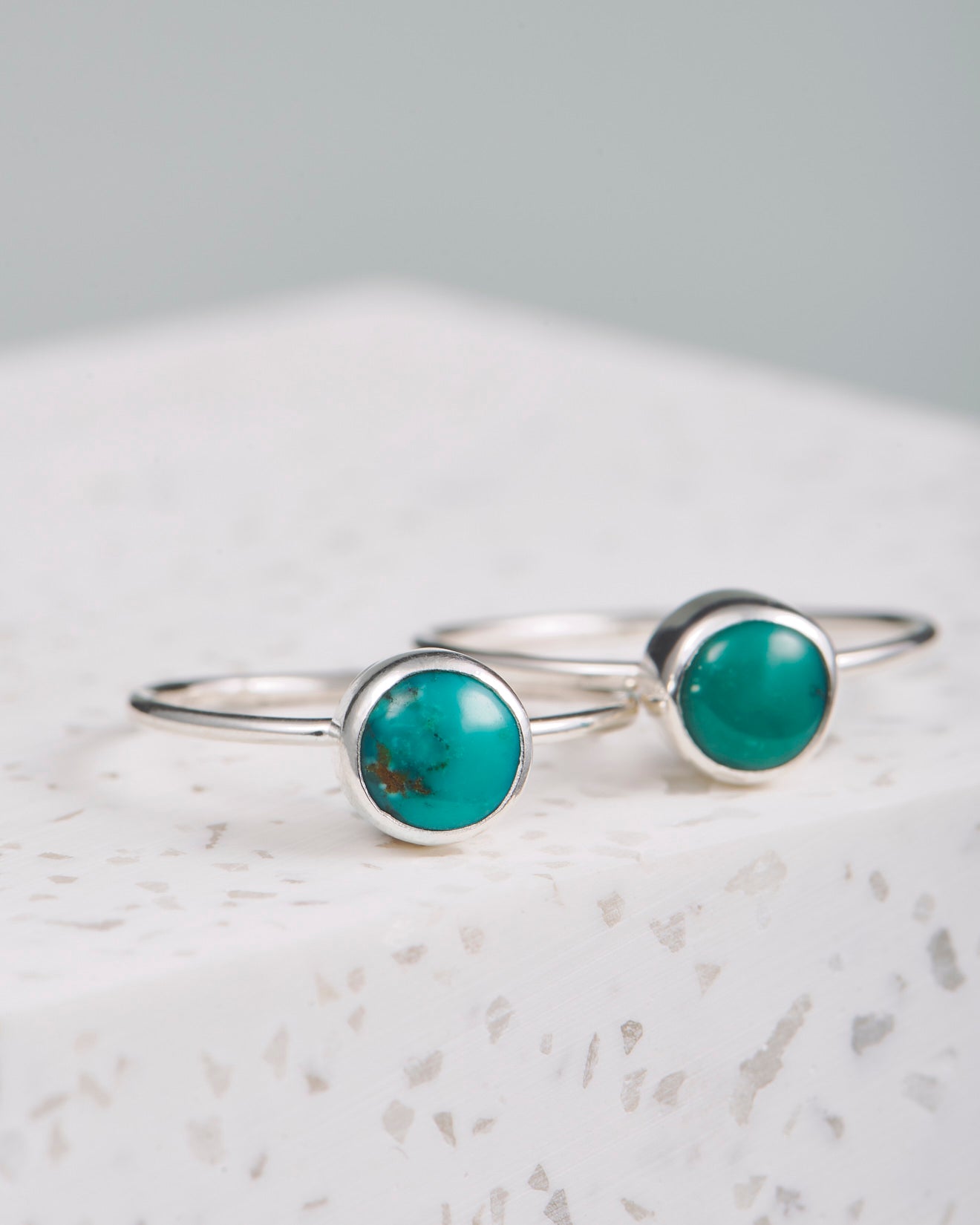Zu sehen sind zwei handgemachte Silberringe, die übereinander gelegt auf einem weißen Marmorblock liegen. Der runde Türkis-Stein zeigt nach vorne. Die Maserungen auf den Steinen sind unterschiedlich. Die Türkis-Steine sind in ein Silberband eingefasst. Alle Ringe sind aus Sterling Silber und somit wasserfest. Der Hintergrund ist grau und unscharf. 