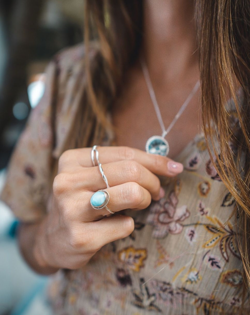 Das Bild zeigt eine Frau, die ein leichtes Oberteil mit Mustern und Blumen darauf trägt. Die Farben sind Beige, Gelb, Braun. Die Frau trägt eine Silberkette und hält den Anhänger davon zwischen den Fingern. An ihrer Hand trägt sie drei Silberringe von Verlan Jewellery. Den Meno Ring mit einem hellblauen Larimar Stein, am Mittelfinger den eingedrehten Sari Ring und am Zeigefinger den Twisted Bingin Ring. 