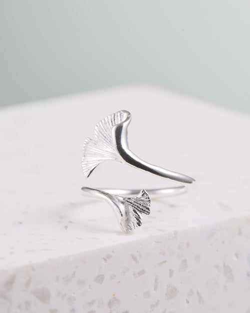 Ein Silberring liegt auf einer weißen Marmorplatte. Der Ring ist geöffnet und somit in der Größe verstellbar. Jedes Ende ist zu einem Ginkgoblatt geformt. Die Enden liegen übereinander, das obere Ginkgoblatt ist etwas größer als das untere. 